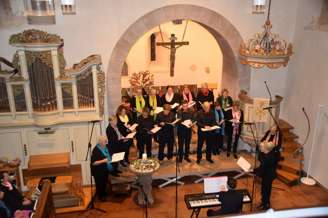 Der Chor singt in der Kirche vor dem Altarbogen, mit Noten in den Händen, links die Orgel, rechts die Chorleiterin am Klavier