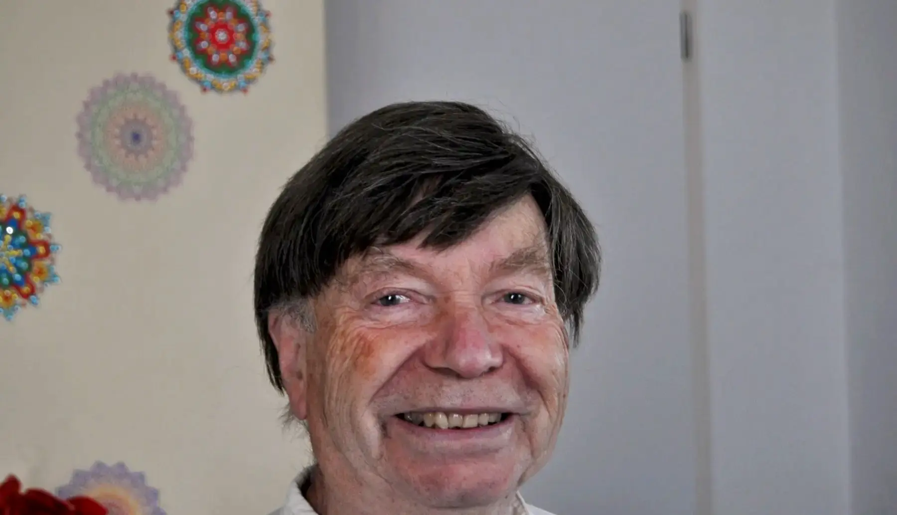 Hans Hermsen, lachend, vor einer cremefarbenen Wand mit bunten Mandala-Bildern