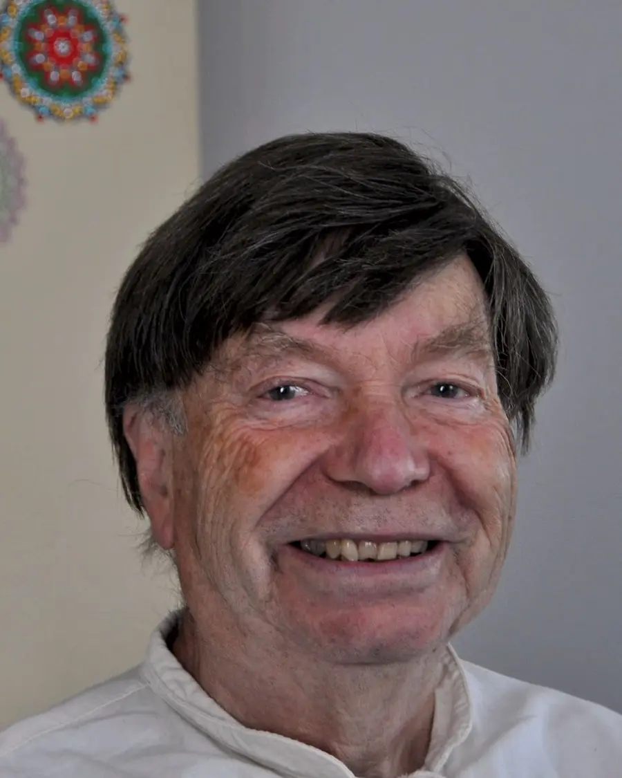 Hans Hermsen, lachend, vor einer cremefarbenen Wand mit bunten Mandala-Bildern