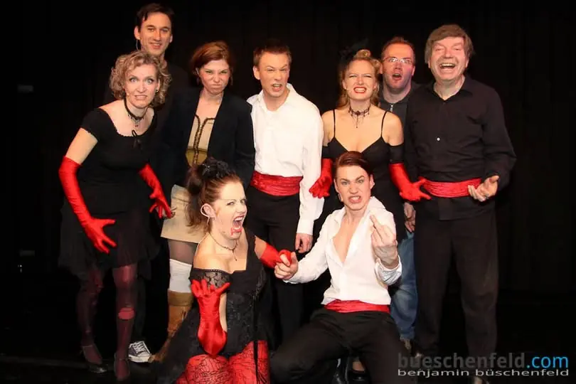 Ensemblefoto von Musical 4 Friends: neun Sängerinnen und Sänger in Schwarz mit roten Handschuhen und Schärpen auf der Bühne, rechts aussen Hans Hermsen