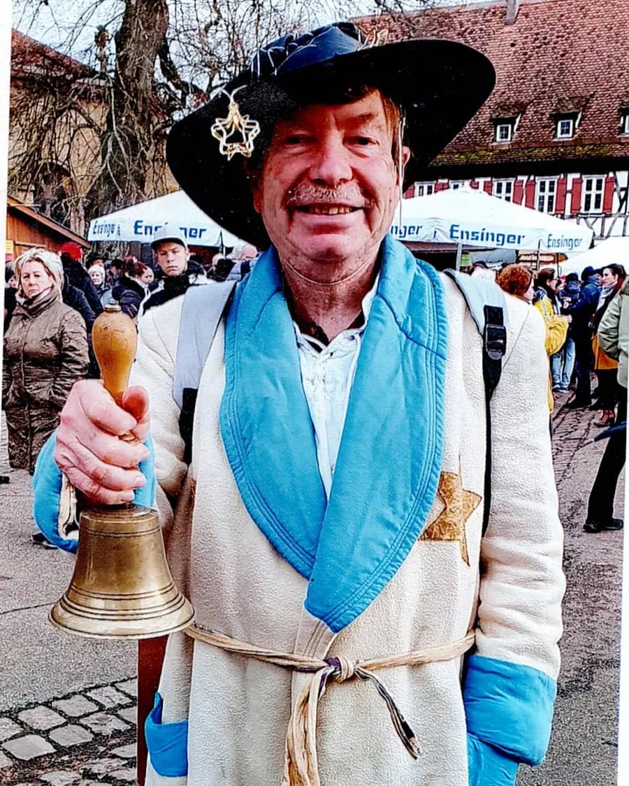 Hans Hermsen in einem langen hellen Mantel mit Mond- und Sternmotiven, eine Messingglocke in der Hand, auf einem Markt