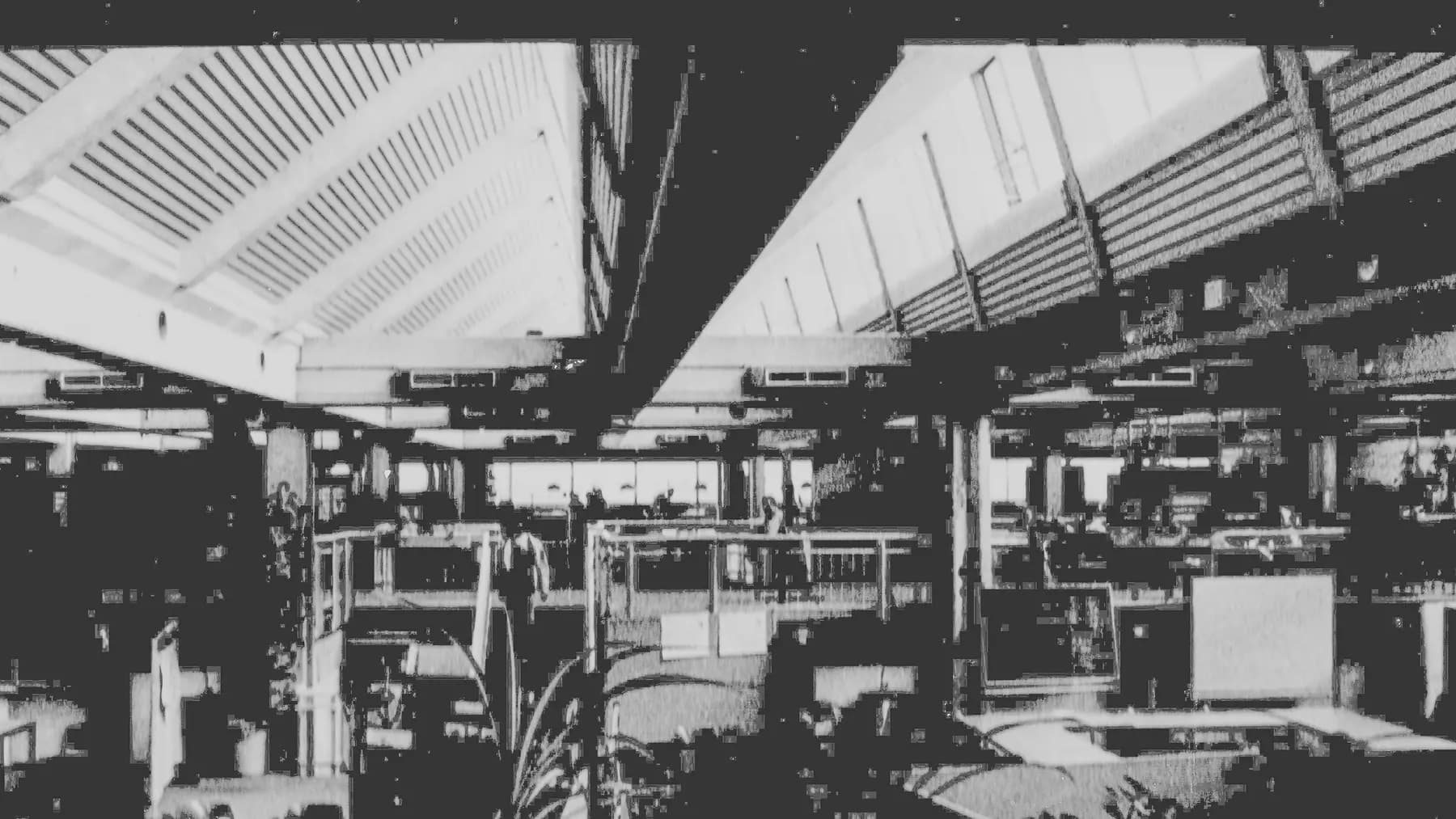 Archivaufnahme in Schwarzweiss: eine weite, offene Halle mit Sheddach, Galerieebene und Arbeitsplätzen ohne Trennwände